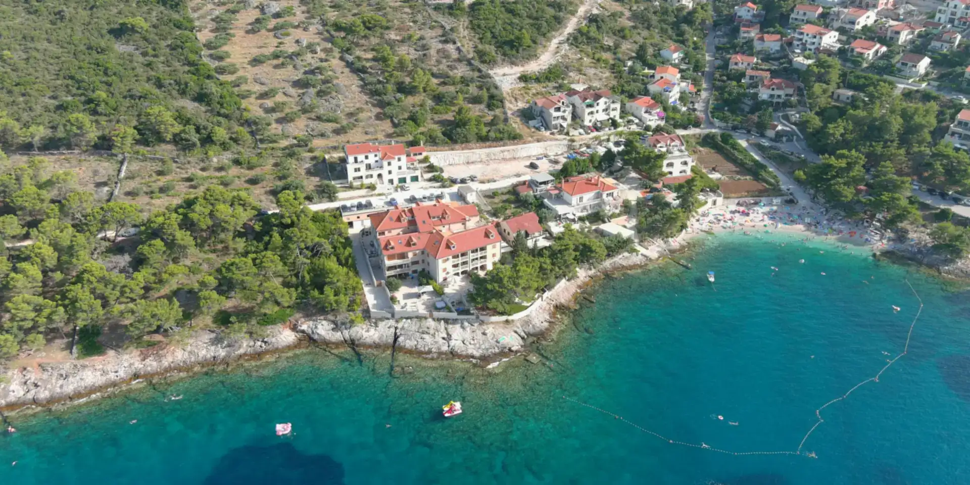 Pogled iz zraka na priobalno naselje na otoku Braču — kamene kuće s crvenim krovovima, uvala s plažom i bistro more